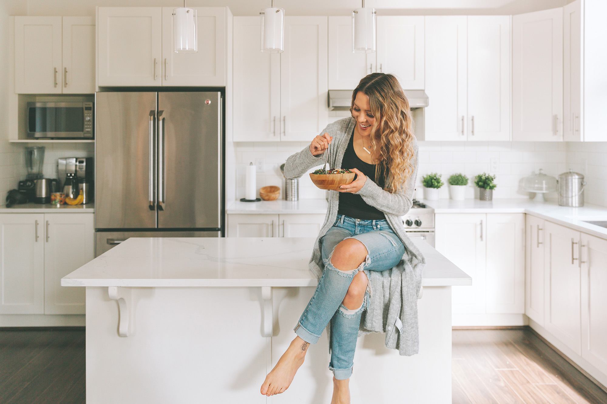Young woman enjoying a healthy salad at a modern kitchen island, featuring white cabinetry and stainless steel appliances, reflecting a stylish and inviting home environment.
