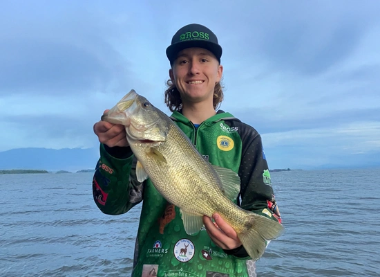 Young angler holding a large bass fish, wearing a green fishing jersey with logos, standing by a lake in a serene outdoor setting, reflecting the community spirit of Hemphill, Texas, and the local High School Bass Club.
