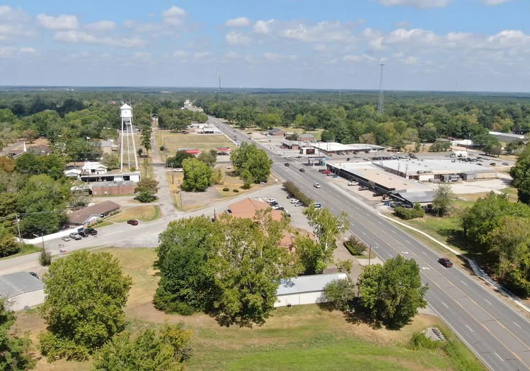 Aerial view of a small town in East Texas featuring a water tower, residential homes, and commercial buildings along a main road, showcasing the community's charm and potential for building custom homes and barndominiums.