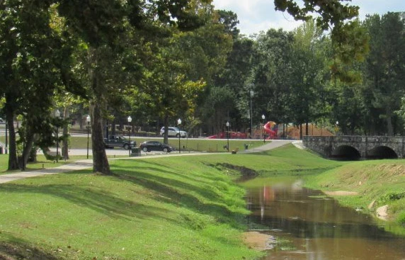 Park landscape in Jasper County, TX, featuring a winding creek, green grass, trees, and a playground in the background, illustrating community spaces near homes and barndominiums.