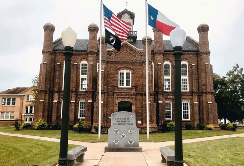 Historic brick building with American and Texas flags, featuring a veterans memorial in front, located in a community area of East Texas.