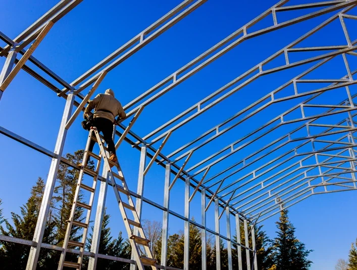Construction worker on ladder assembling steel frame for custom metal building under clear blue sky, showcasing durable design for storage and workshop needs.