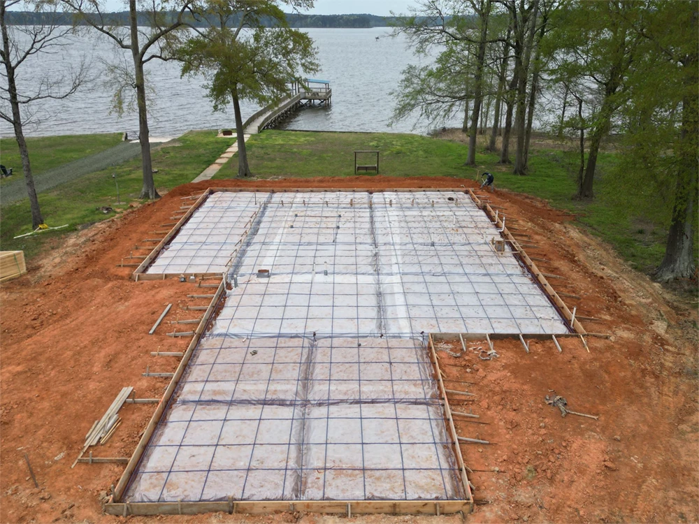 Lakeside home construction site with a concrete foundation framework, surrounded by trees and water, illustrating custom home blueprints for lake homes.