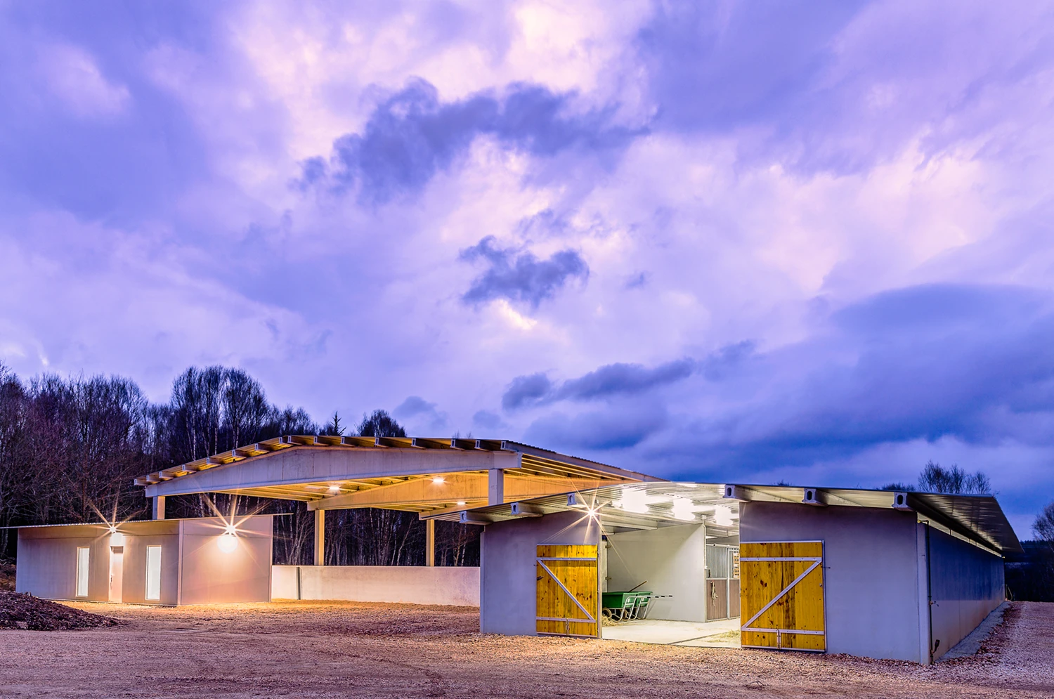 Custom metal building for horse riding school, featuring steel structure, wooden doors, and illuminated interior, set against a cloudy sky in East Texas.