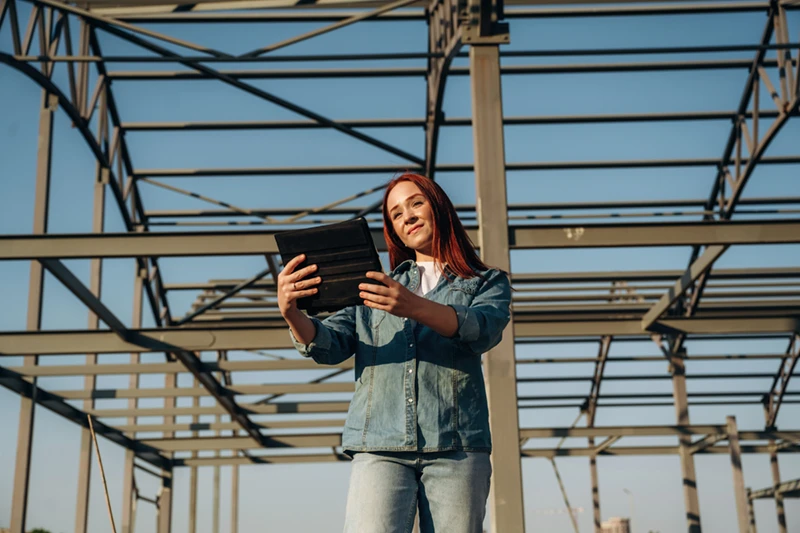 Woman holding a tablet and smiling, standing near an unfinished metal building frame in East Texas, showcasing construction progress for custom steel buildings.